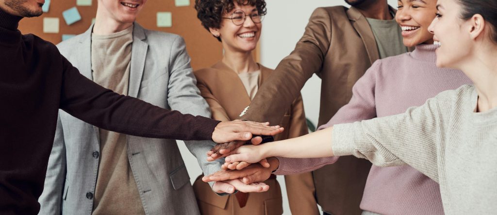 People of different genders and ethnicity are reaching towards each other to place their outstretched hands on top of each other in a sign of unity and celebration.