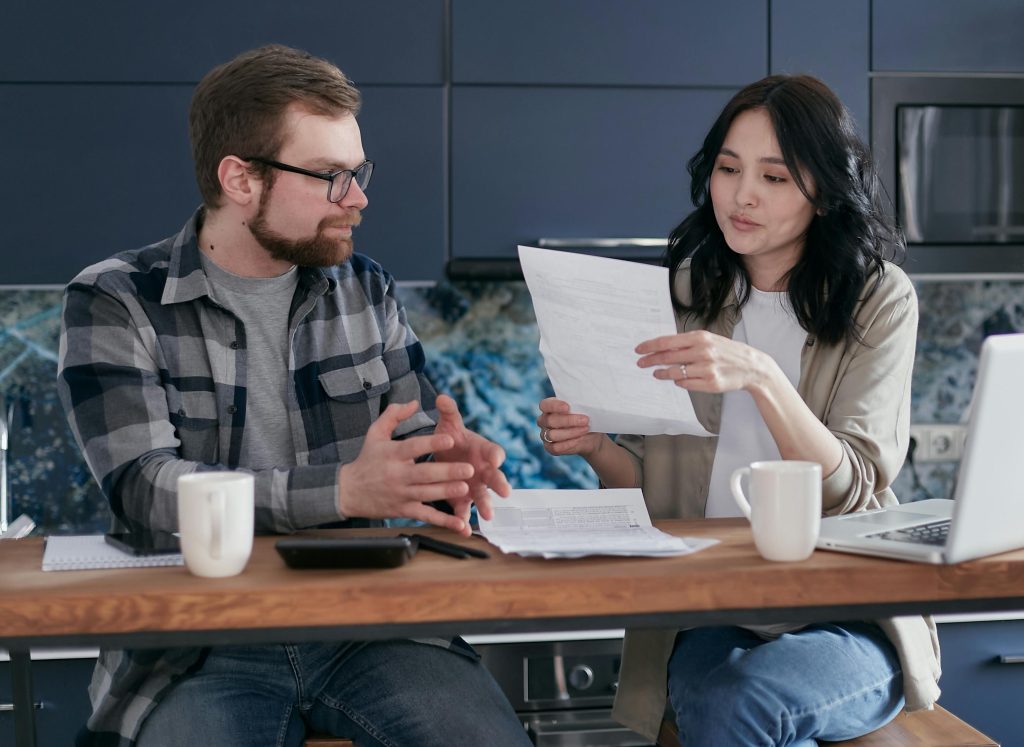 A man and a woman sitting at a table reviewing paperwork together, discussing documents with a laptop and calculator nearby.